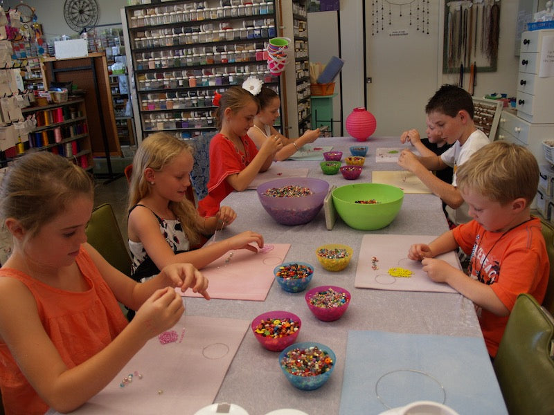 Table showing lots of children doing a beading workshop with a shop background with jars of beads and cord reels