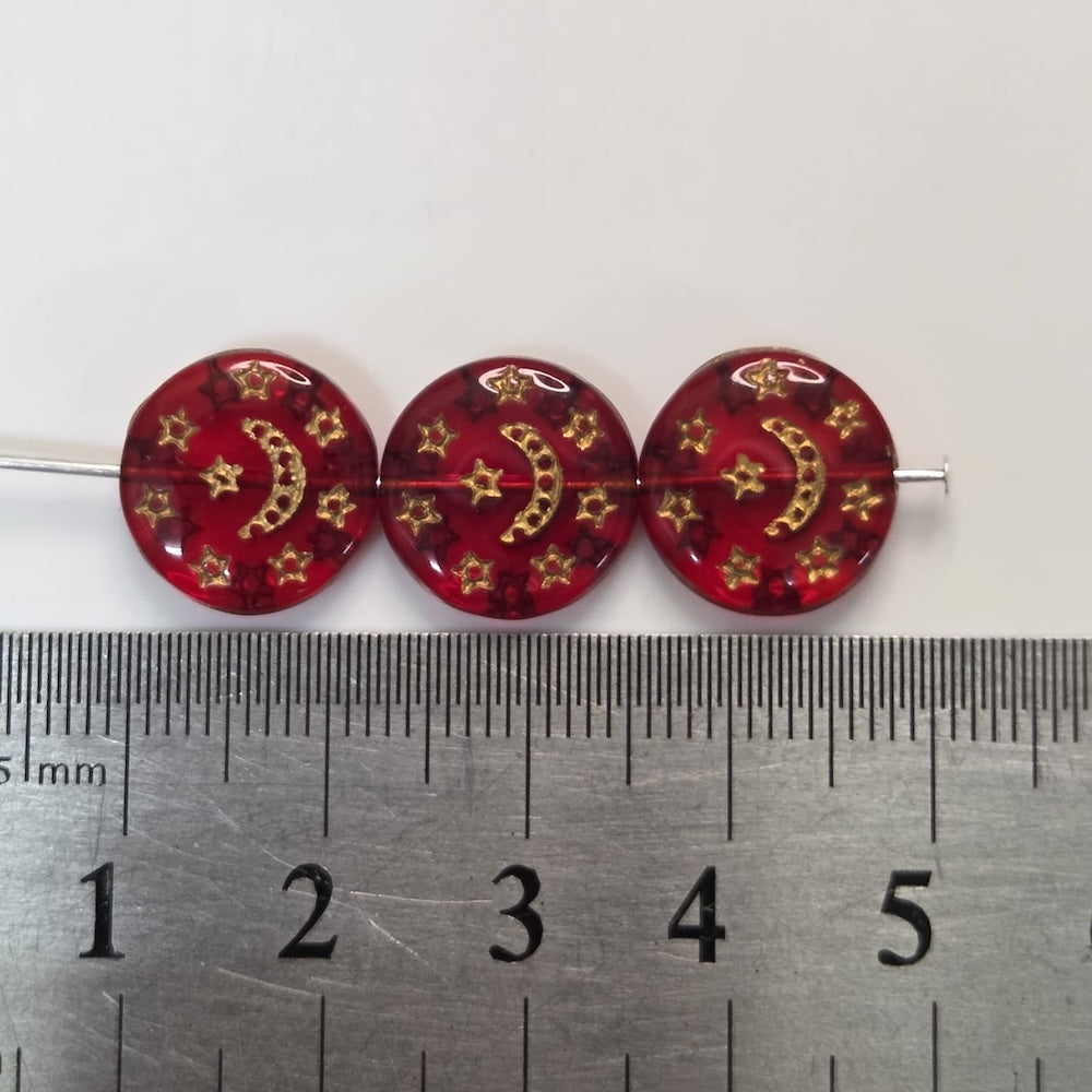 Three red beads with gold patterns on a metal ruler for scale.