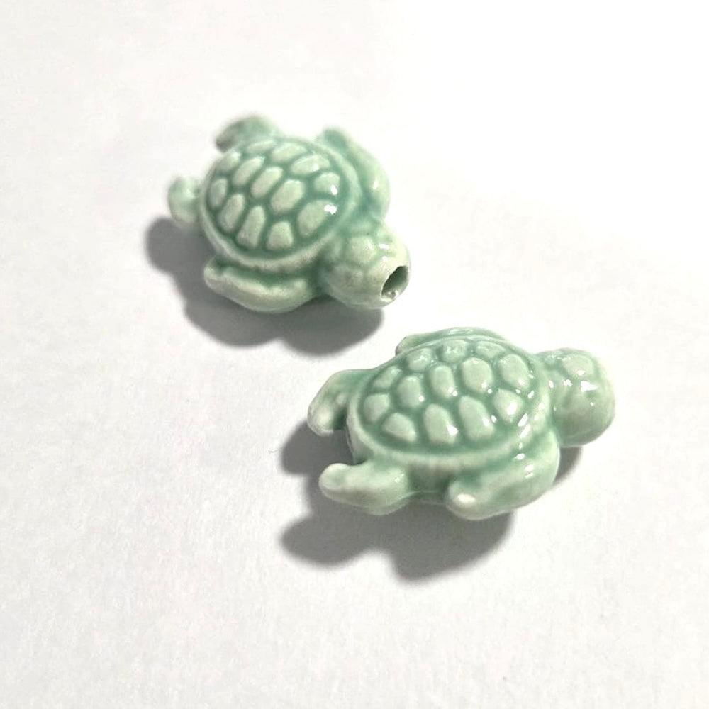 Two light green turtle-shaped porcelain ceramic beads on a white background