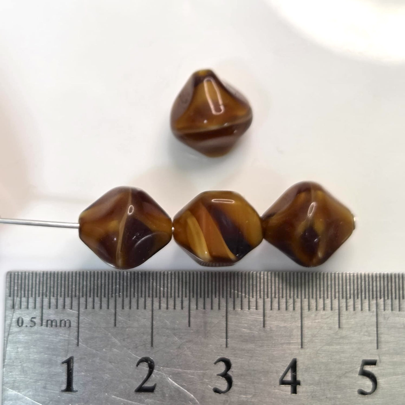 Four brown and beige patterned beads on a ruler for size reference