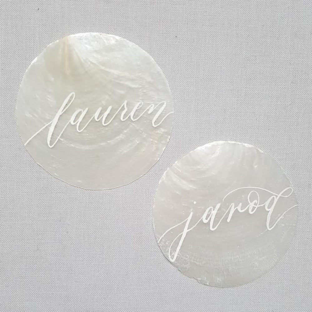 white, round capiz shell disc placed on a wooden surface used for wedding name place settings, bonbonniere, artwork and craft items displayed on a wooden surface displaying names in calligraphy