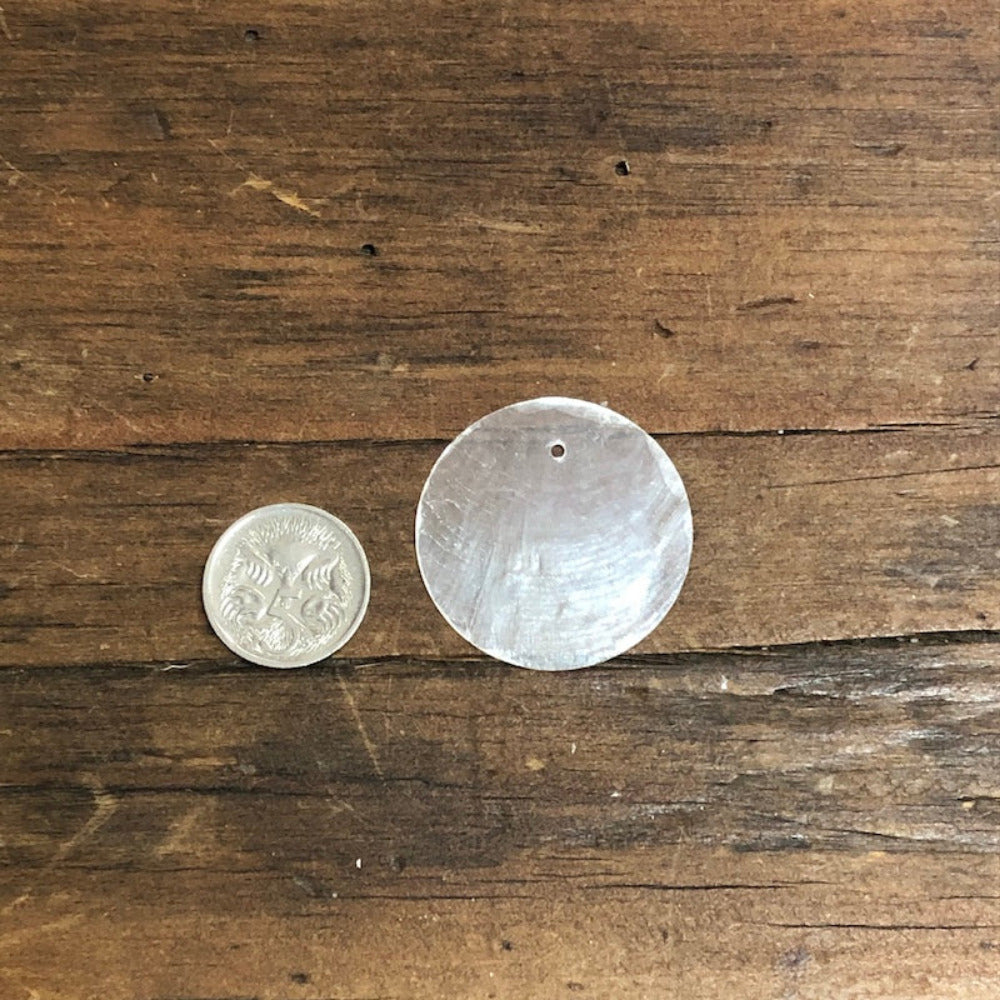 A 3cm round white Capiz shell with a pre-drilled hole, displayed next to a coin for size comparison on a wooden surface.