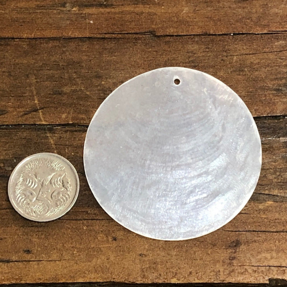A white, circular capiz shell with a pre-drilled hole, displayed next to an coin for size comparison on a wooden surface.