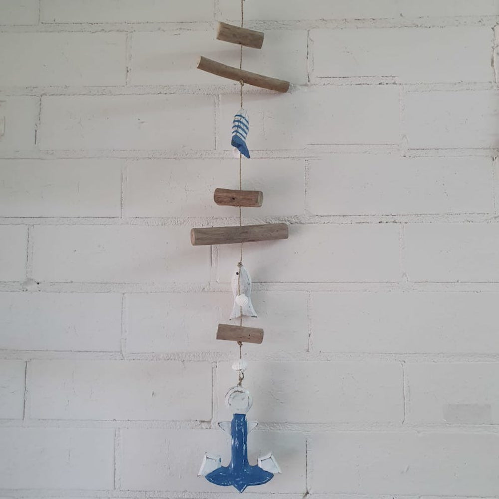 A hanging decoration featuring a series of wood cutouts, beads, and a anchor-shaped charm, strung together on natural seagrass string against a white brick wall.