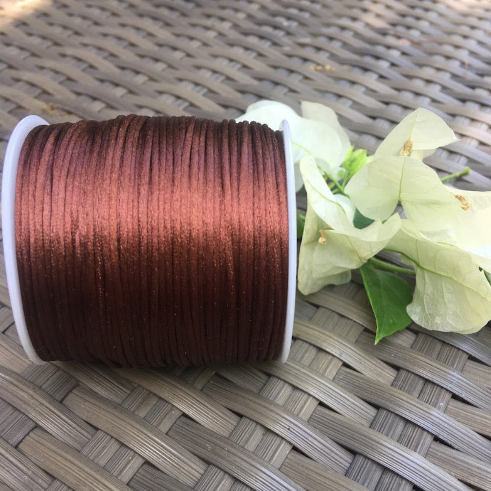 A spool of brown rat tail cord for crafting, displayed on a woven surface with a white flower beside it.