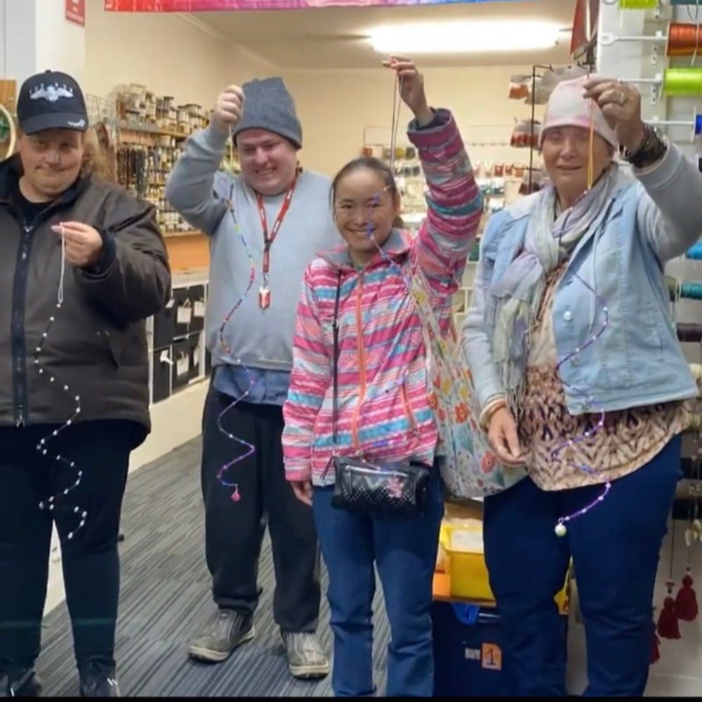 A group of four adults with disability, workshop participants, standing in a bead store displaying their handmade beaded crafts.