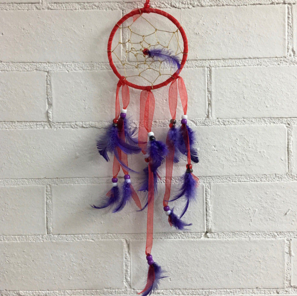 A dreamcatcher with a hoop, beads, and feathers, primarily in purple and gold colors, hanging against a white brick wall.