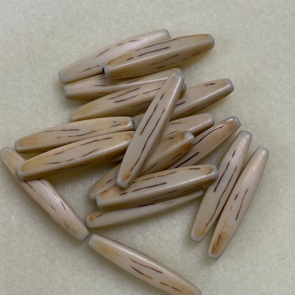 A collection of light brown, hair pipe shaped beads with a threading hole arranged in a pile on a beige background.
