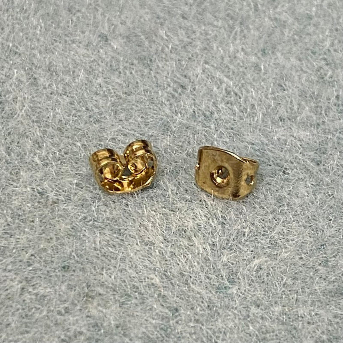 A pair of gold-colored butterfly stud backs for earrings placed on a grey surface.