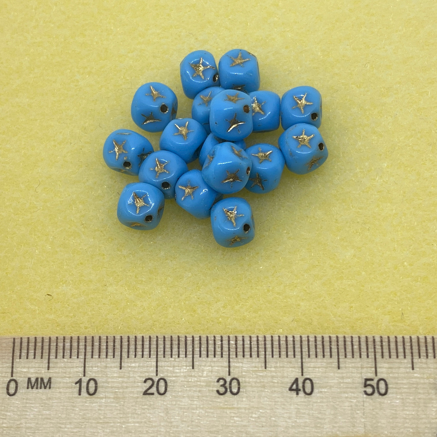 a collection light blue cube-shaped beads with gold stars etching with a ruler to show scale on a yellow background
