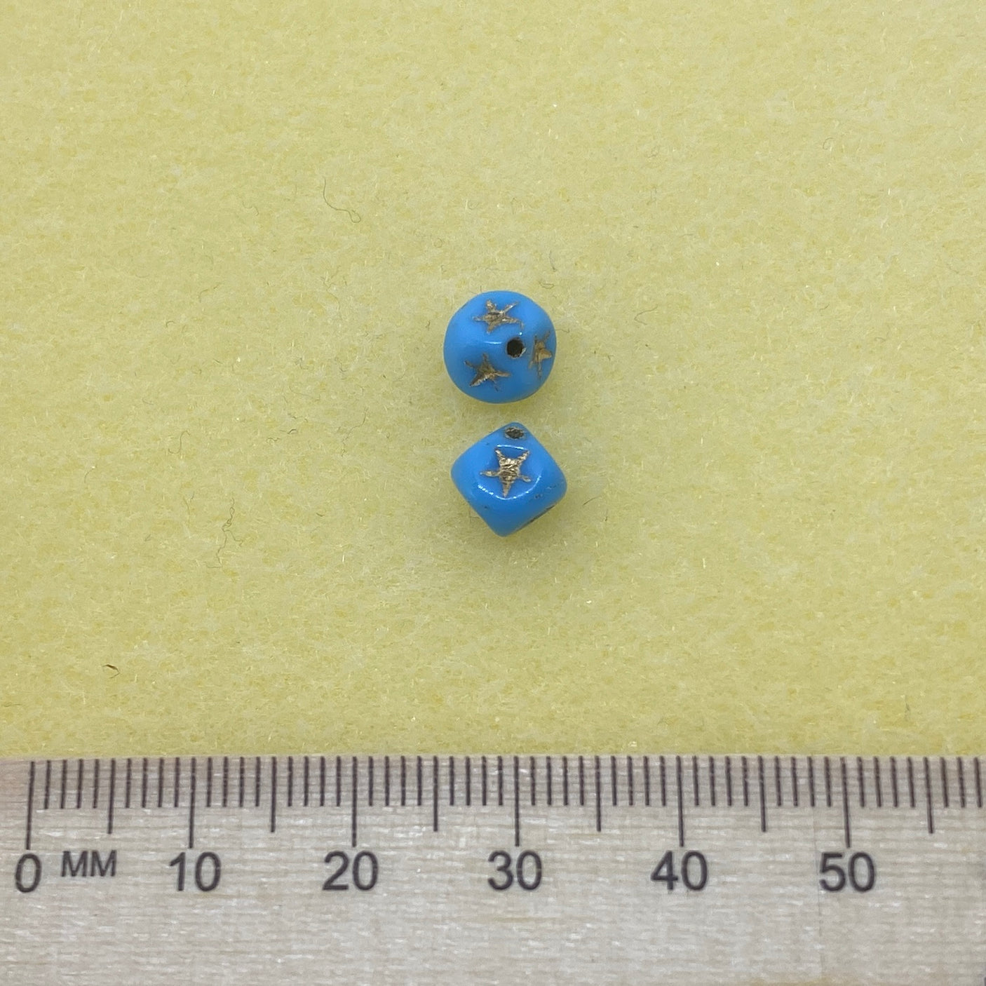 two light blue cube-shaped beads with gold stars etching with a ruler to show scale on a yellow background