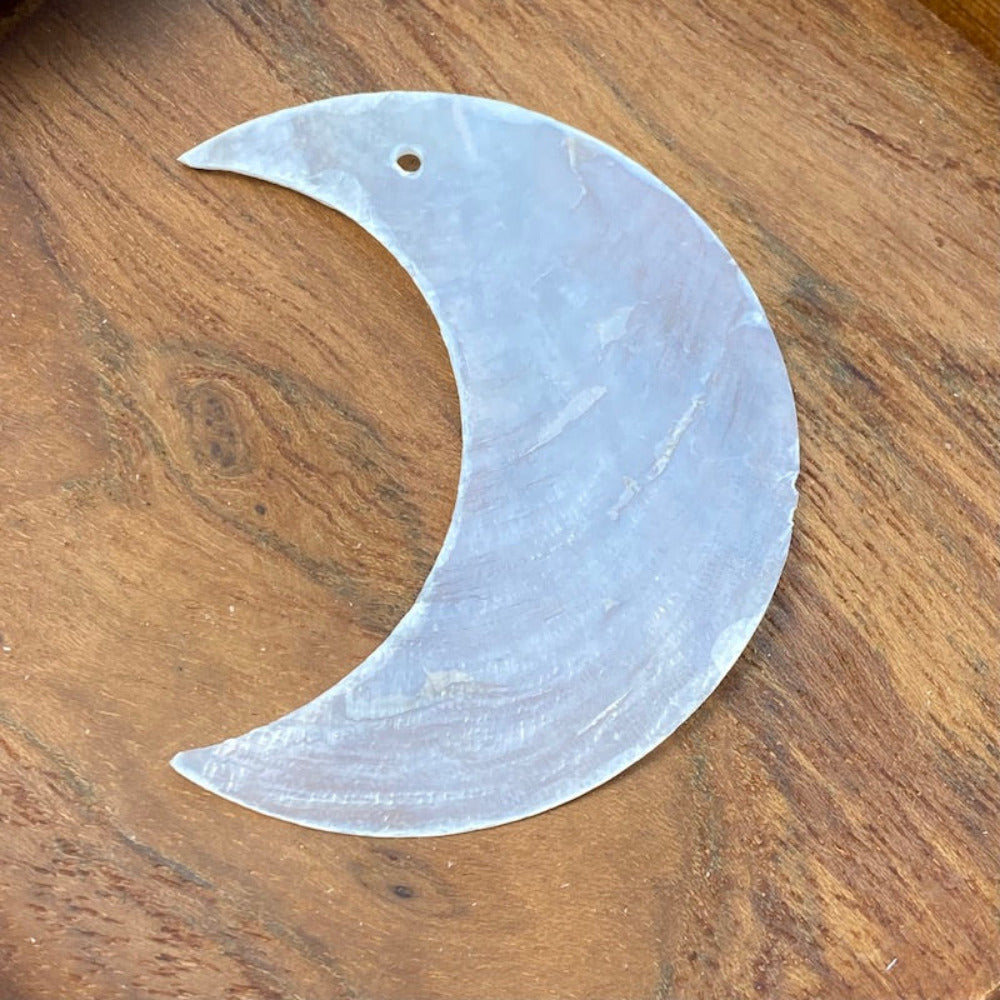 A pre-drilled white Capiz shell in the shape of a crescent moon laid on a wooden surface.