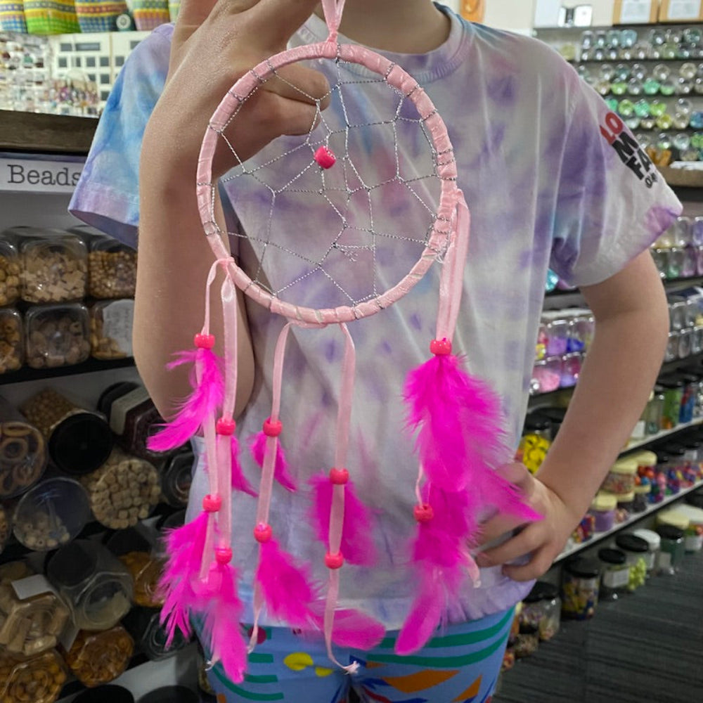 A child holding up a pink dream catcher they have made, featuring pink feathers and beads against a store background.