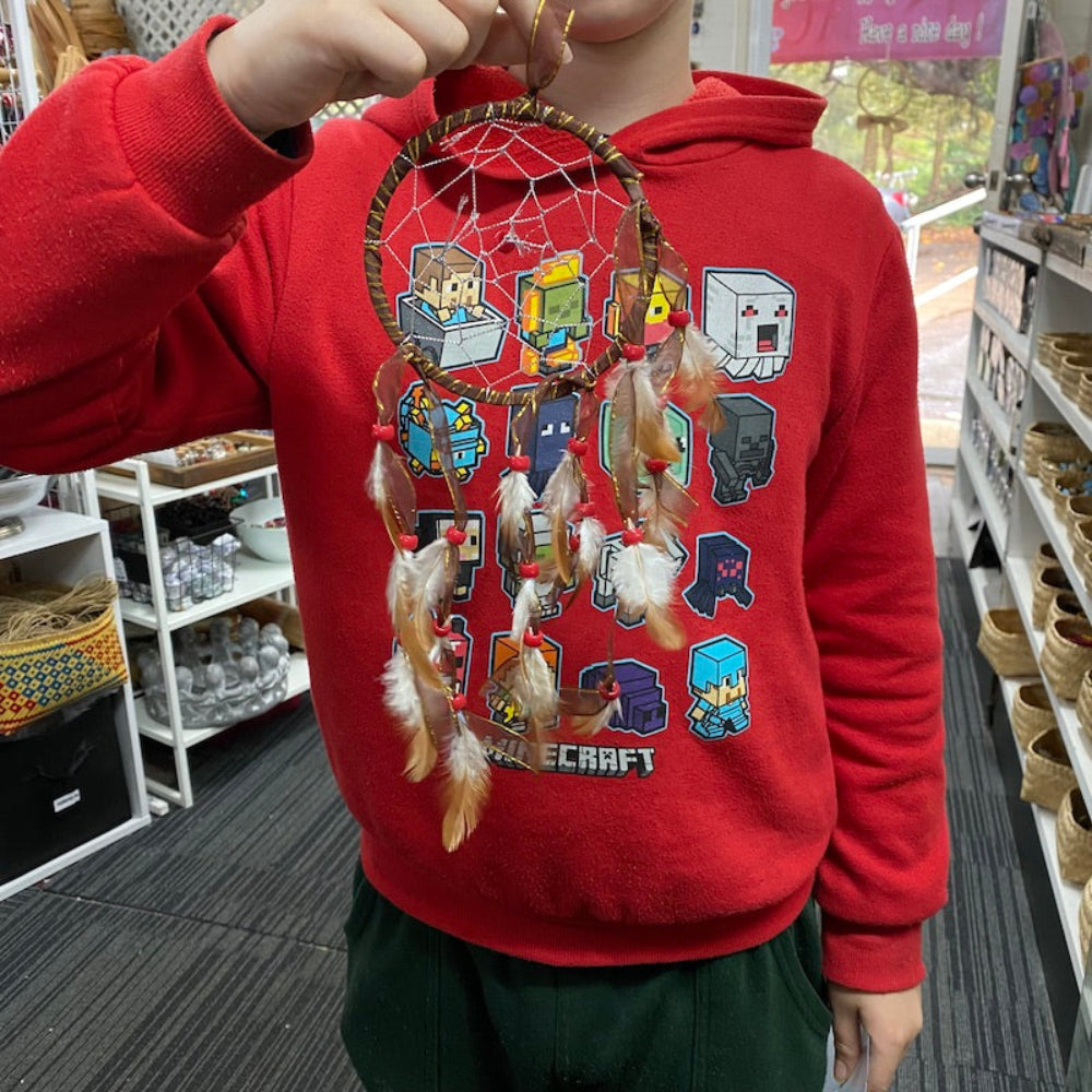 A child in a red shirt holding up a brown natural colour dream catcher they have made, featuring natural brown and white feathers and beads against a store background.