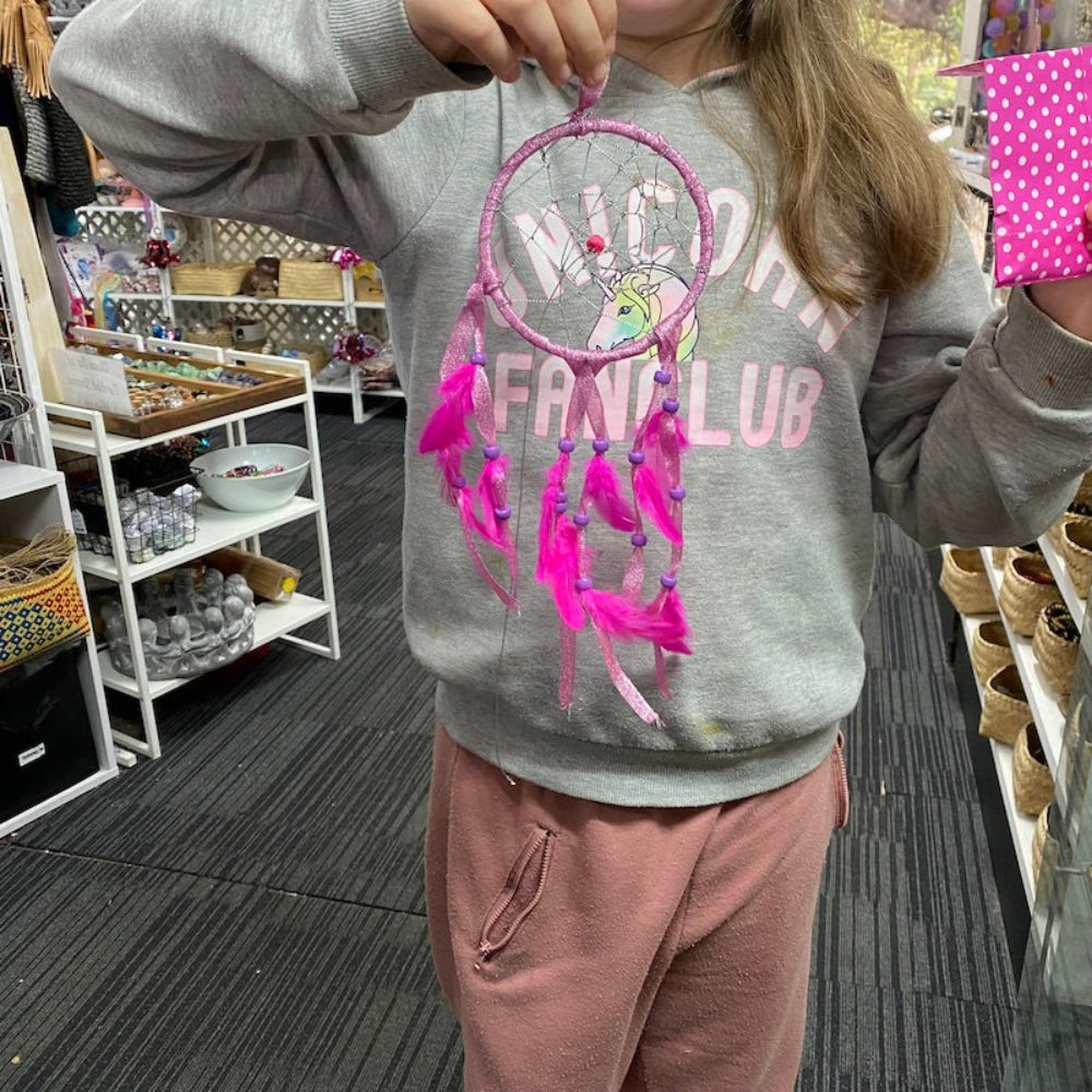 A child holding up a pink dream catcher they have made, featuring pink feathers and beads against a store background.