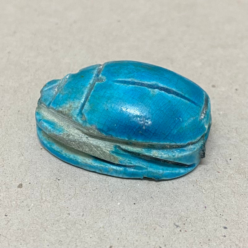 A vintage Egyptian scarab bead in blue color, displaying carving features in a close up photo on a grey background