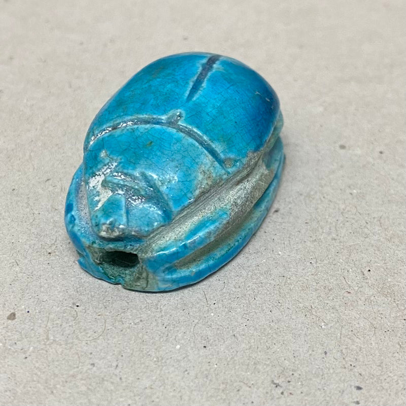 A vintage Egyptian scarab bead in blue color, displaying carving features in a close up photo on a grey background