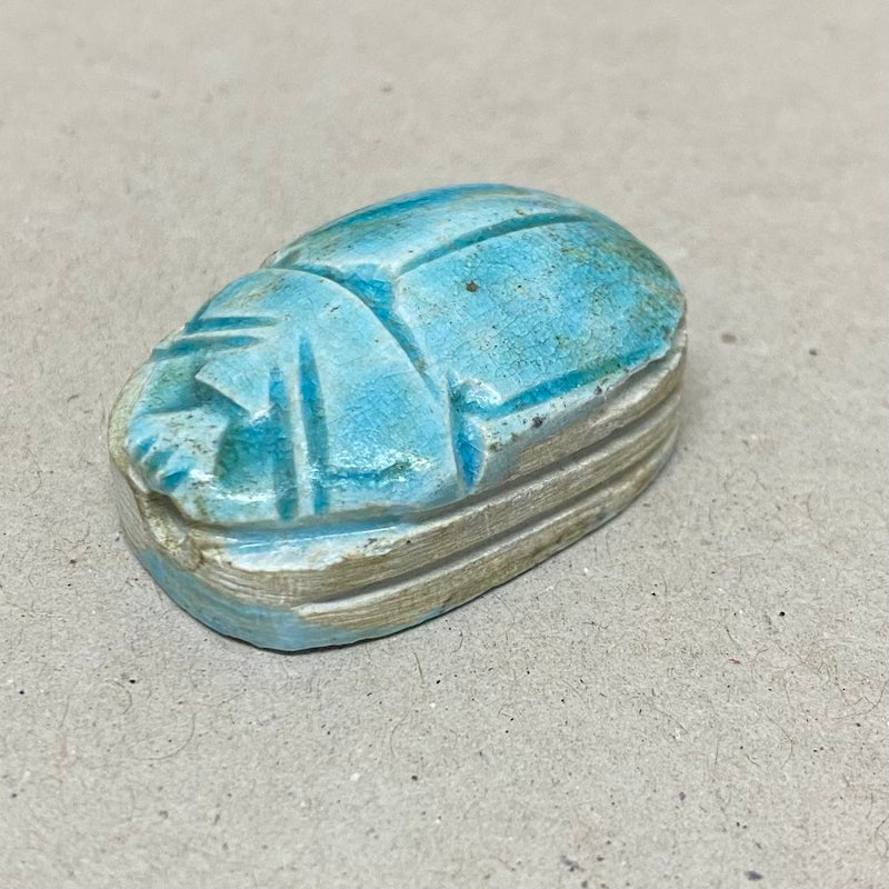 A vintage Egyptian scarab bead in blue color, displaying carving features in a close up photo on a grey background
