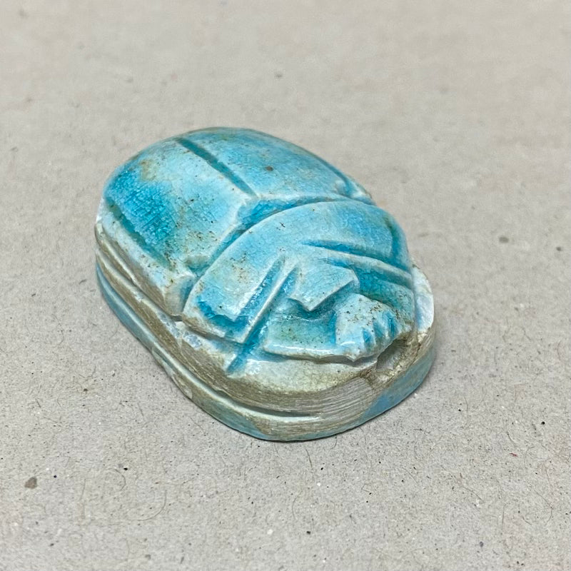 A vintage Egyptian scarab bead in blue color, displaying carving features in a close up photo on a grey background