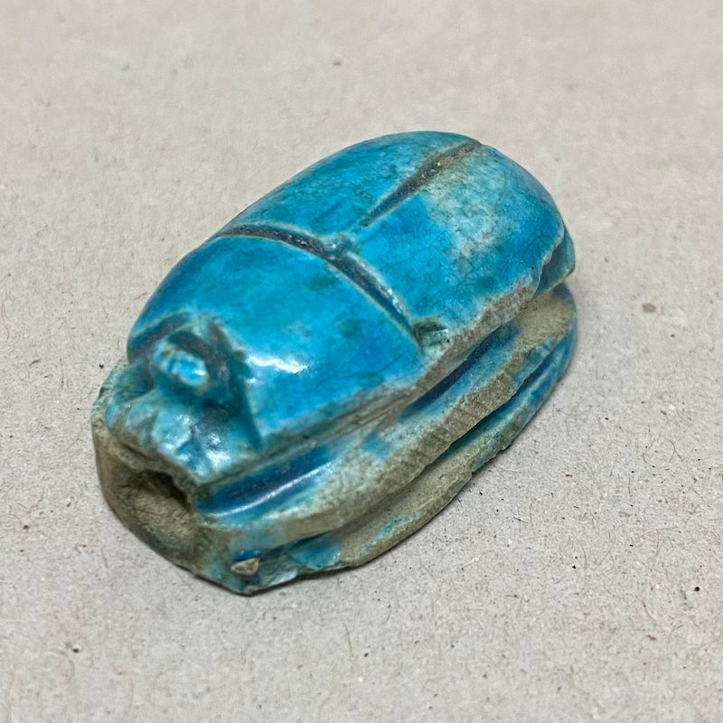 A vintage Egyptian scarab bead in blue color, displaying carving features in a close up photo on a grey background