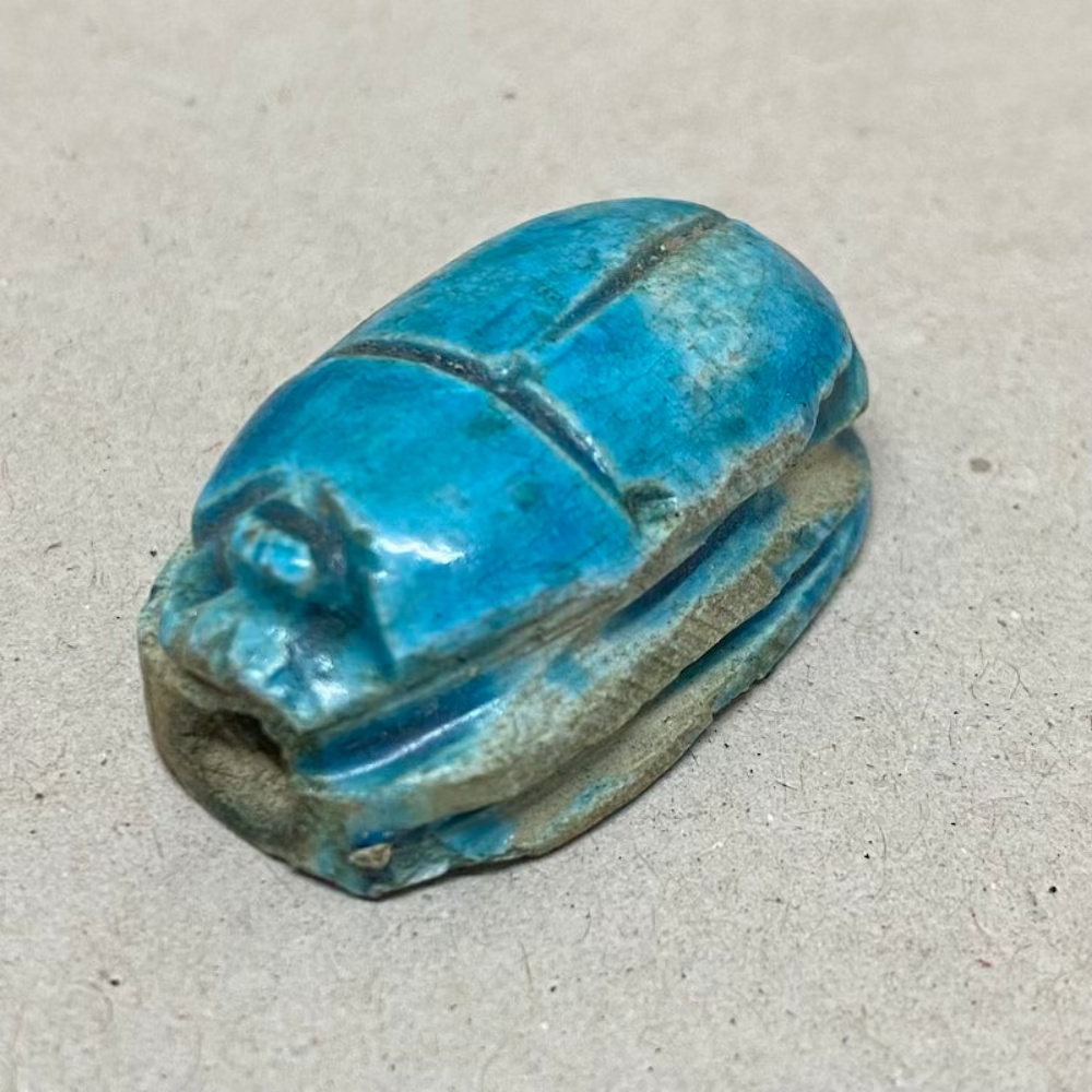 A vintage Egyptian scarab bead in blue color, displaying carving features in a close up photo on a grey background