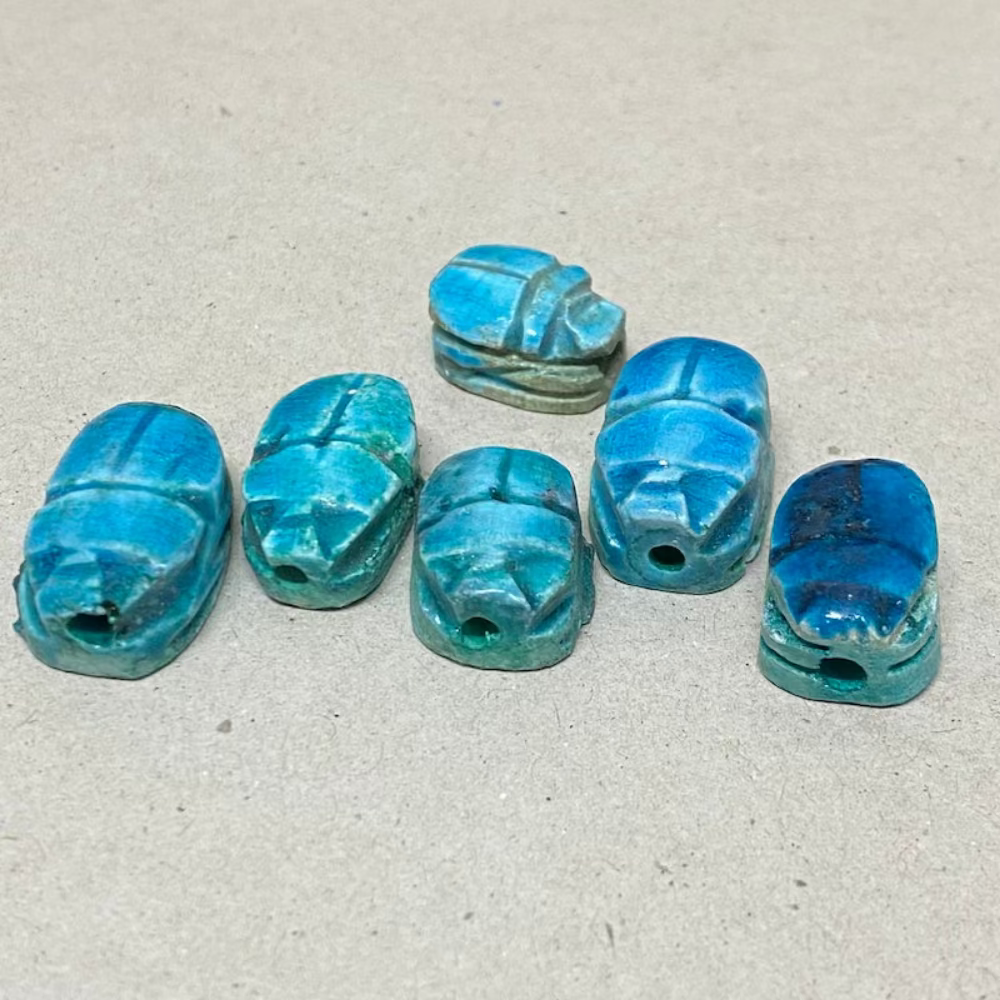A line of vintage Egyptian scarab beads in blue, varying slightly in size and shape, displayed against a neutral background showing the threading holes