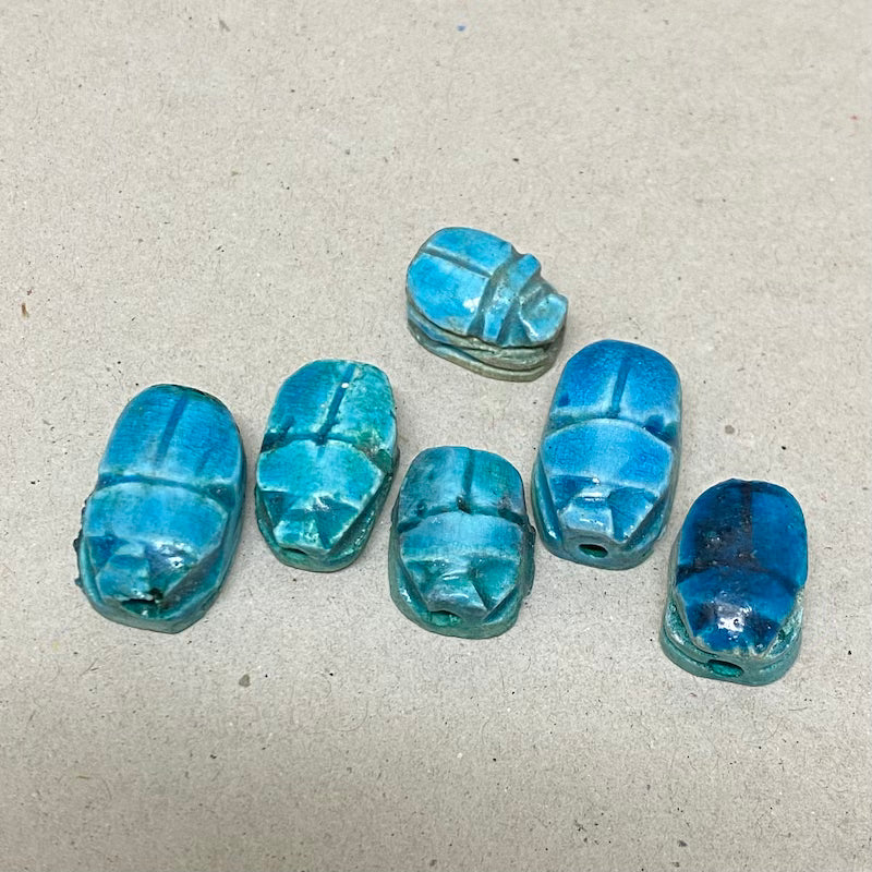 A collection of vintage Egyptian scarab beads in blue, varying slightly in size and shape, displayed against a neutral background