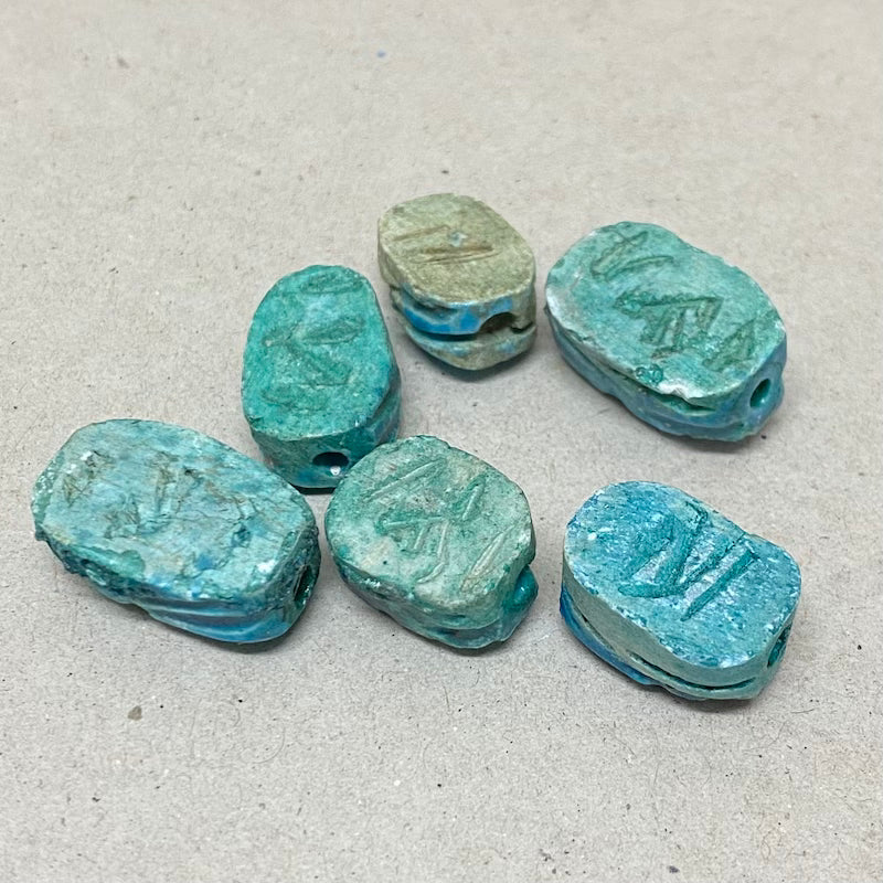 A collection of vintage Egyptian scarab beads in blue, varying slightly in size and shape, displayed against a neutral background showing details of the reverse underneath side featuring hieroglyphics markings