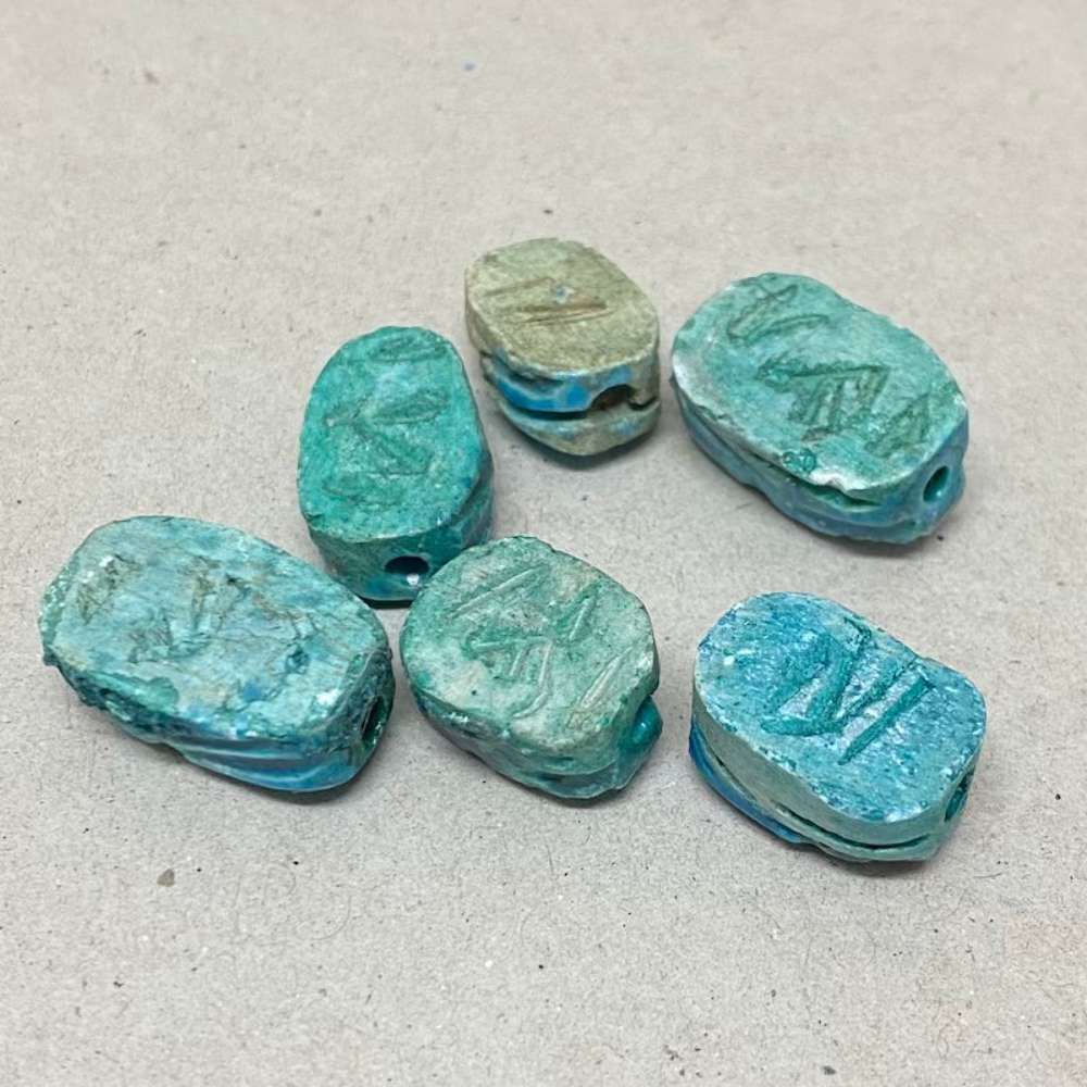 A collection of vintage Egyptian scarab beads in blue, varying slightly in size and shape, displayed against a neutral background showing details of the reverse underneath side featuring hieroglyphics markings