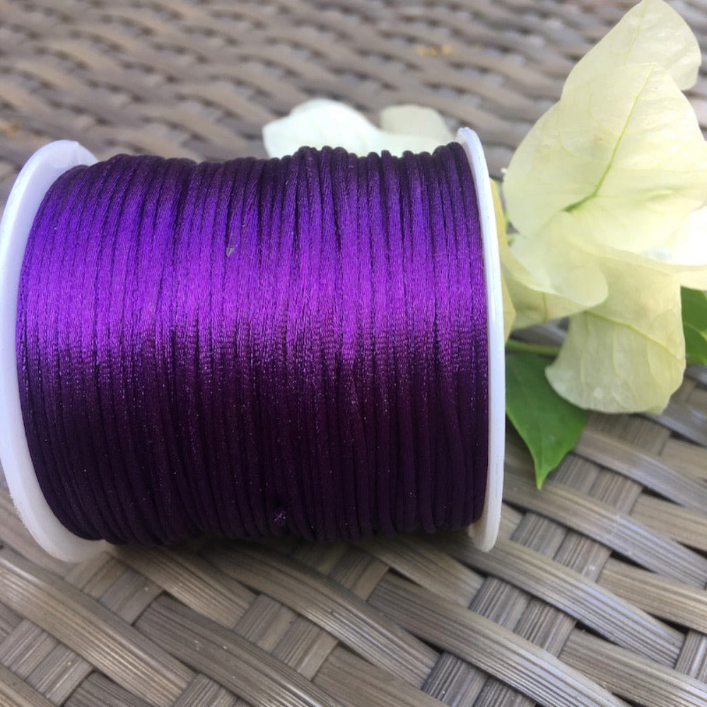 A spool of purple rat tail cord used for crafting, displayed alongside some white flowers and green leaves.