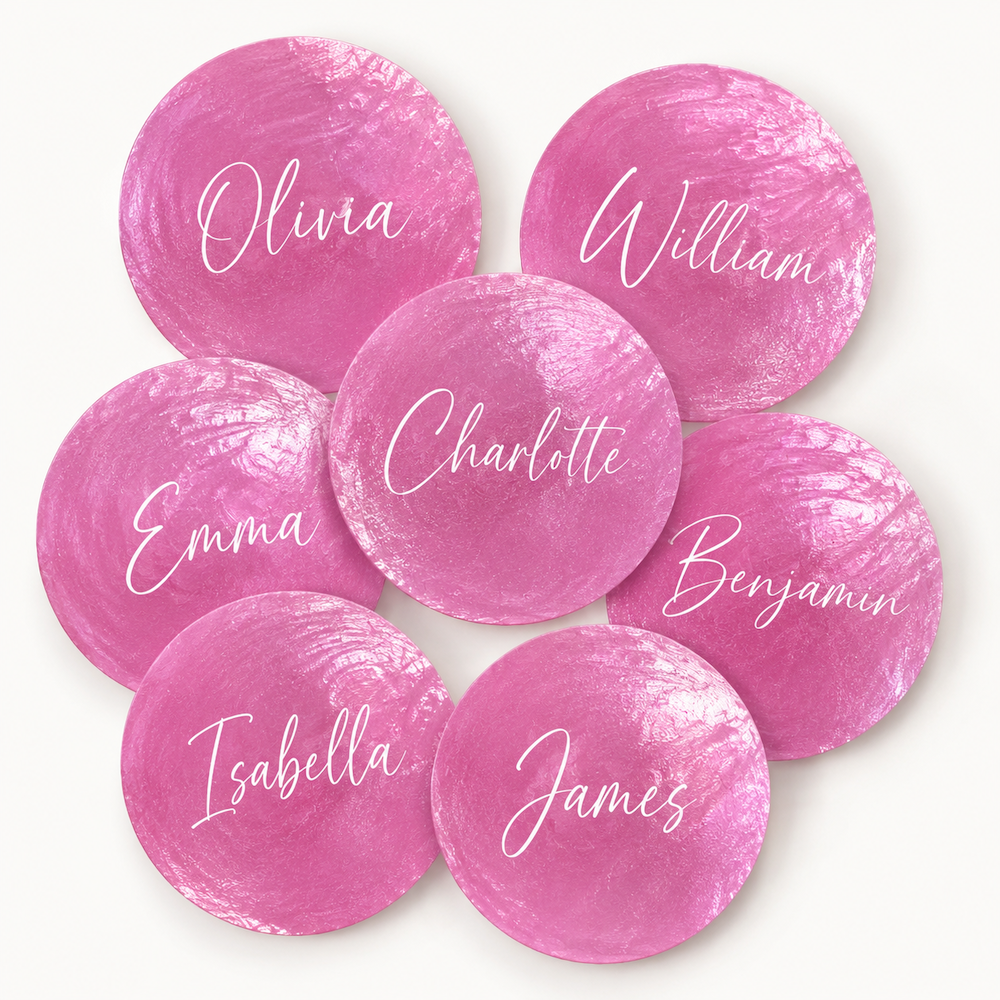 Seven round pink watercolor-style shell place settings with white script names (Olivia, William, Emma, Charlotte, Benjamin, Isabella, and James) arranged in a circle on a light off-white background.