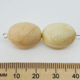 Two beige beads with a textured surface next to a wooden ruler for scale on a white background