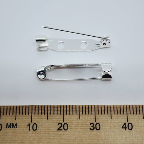 2 gold brooch back fittings on a white surface with a wood ruler to show size