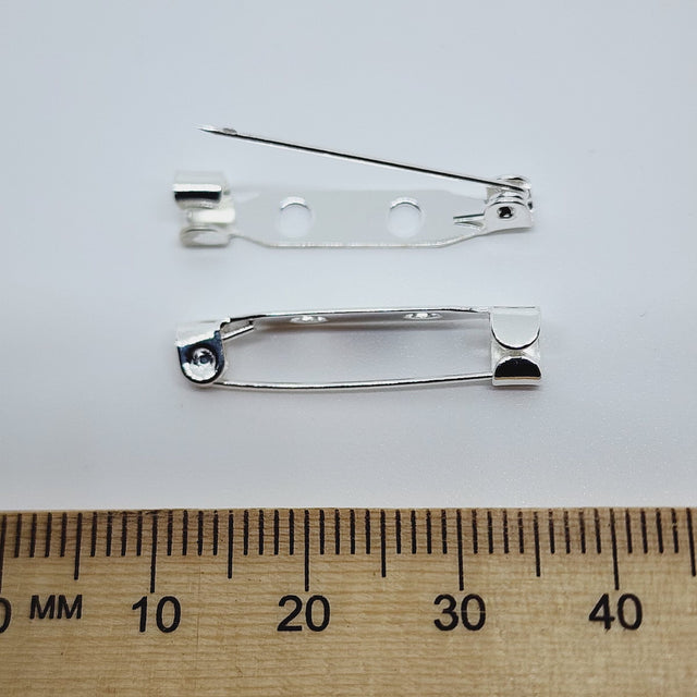 2 gold brooch back fittings on a white surface with a wood ruler to show size
