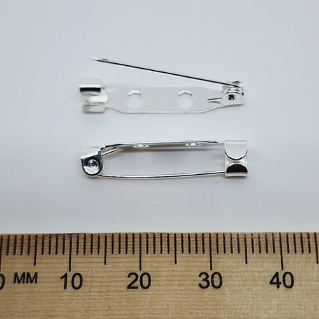 2 silver brooch back fittings on a white surface with a wood ruler to show size