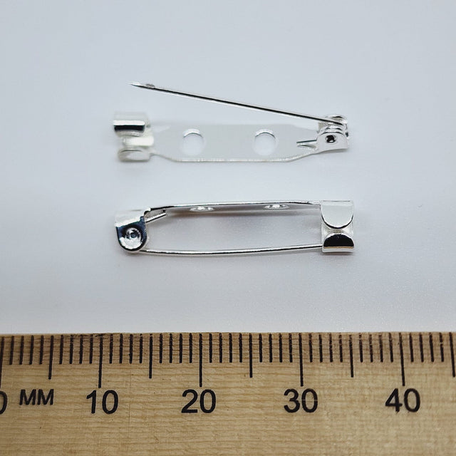 2 silver brooch back fittings on a white surface with a wood ruler to show size