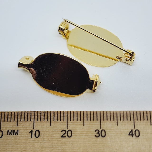 2 gold brooch back fittings on a white surface with a wood ruler to show size