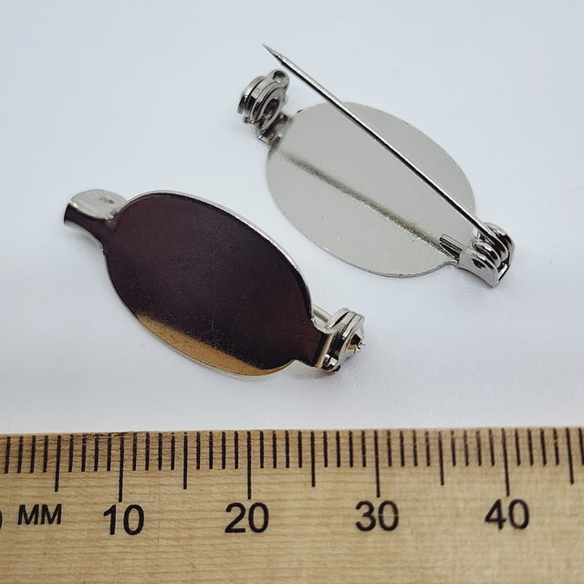 2 silver oval brooch back fittings on a white surface with a wood ruler to show size