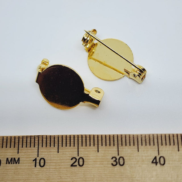 2 gold brooch back fittings on a white surface with a wood ruler to show size