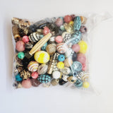 A clear plastic bag filled with assorted decorative beads and charms in many colors, including pink, yellow, turquoise, blue, black, white, and metallic gold, photographed on a plain white background.