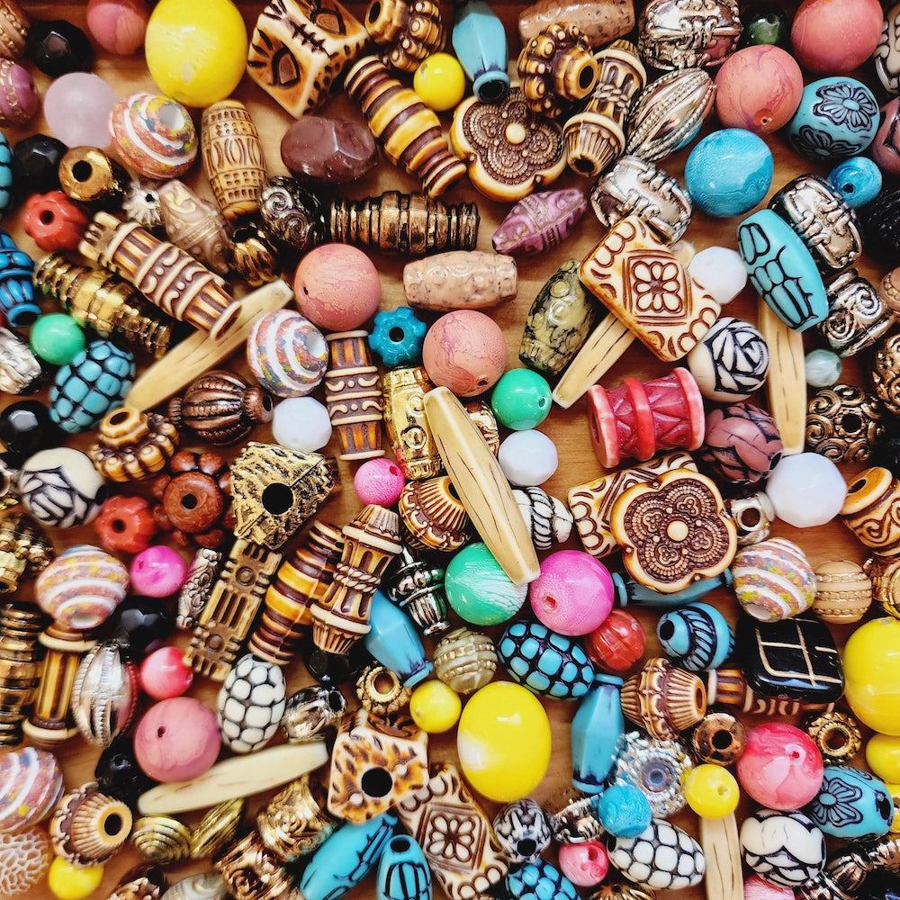 A dense, top-down pile of assorted decorative craft beads in a wide variety of colors, patterns, and shapes, including carved wooden beads, turquoise-blue beads, pink and yellow oval beads, and striped accents, all resting on a light brown wooden surface.