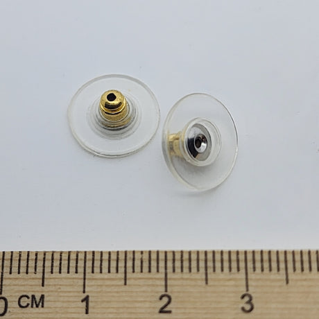 Two gold-colored plastic disc ear stud backs with a central bullet fitting, displayed next to a wooden ruler for scale.