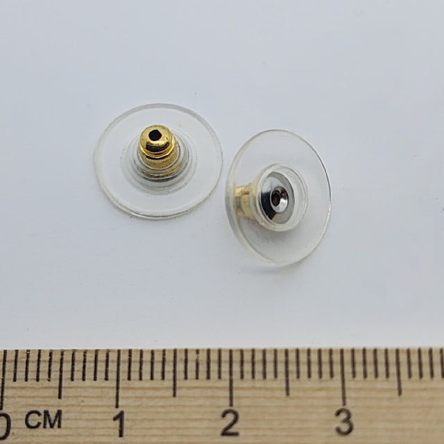 Two gold-colored plastic disc ear stud backs with a central bullet fitting, displayed next to a wooden ruler for scale.