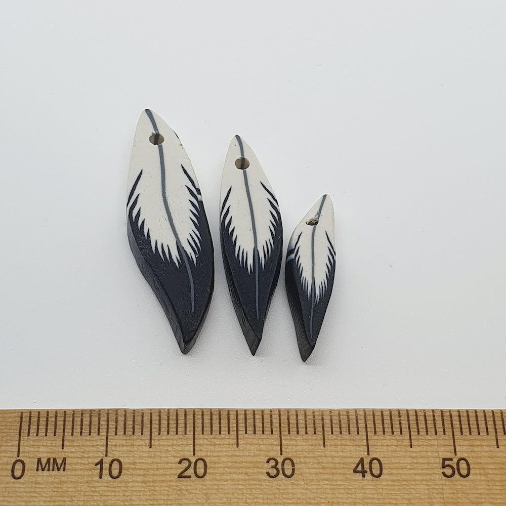 Three feather-shaped beads on a white background with a ruler for scale.