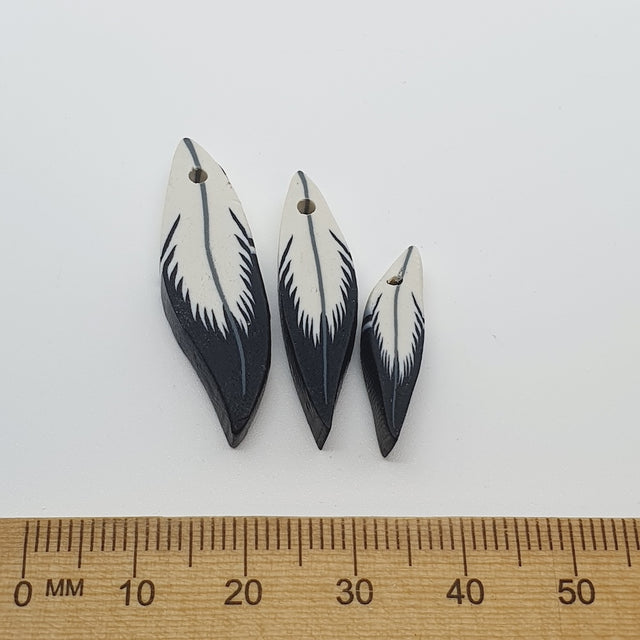 Three feather-shaped beads on a white background with a ruler for scale.