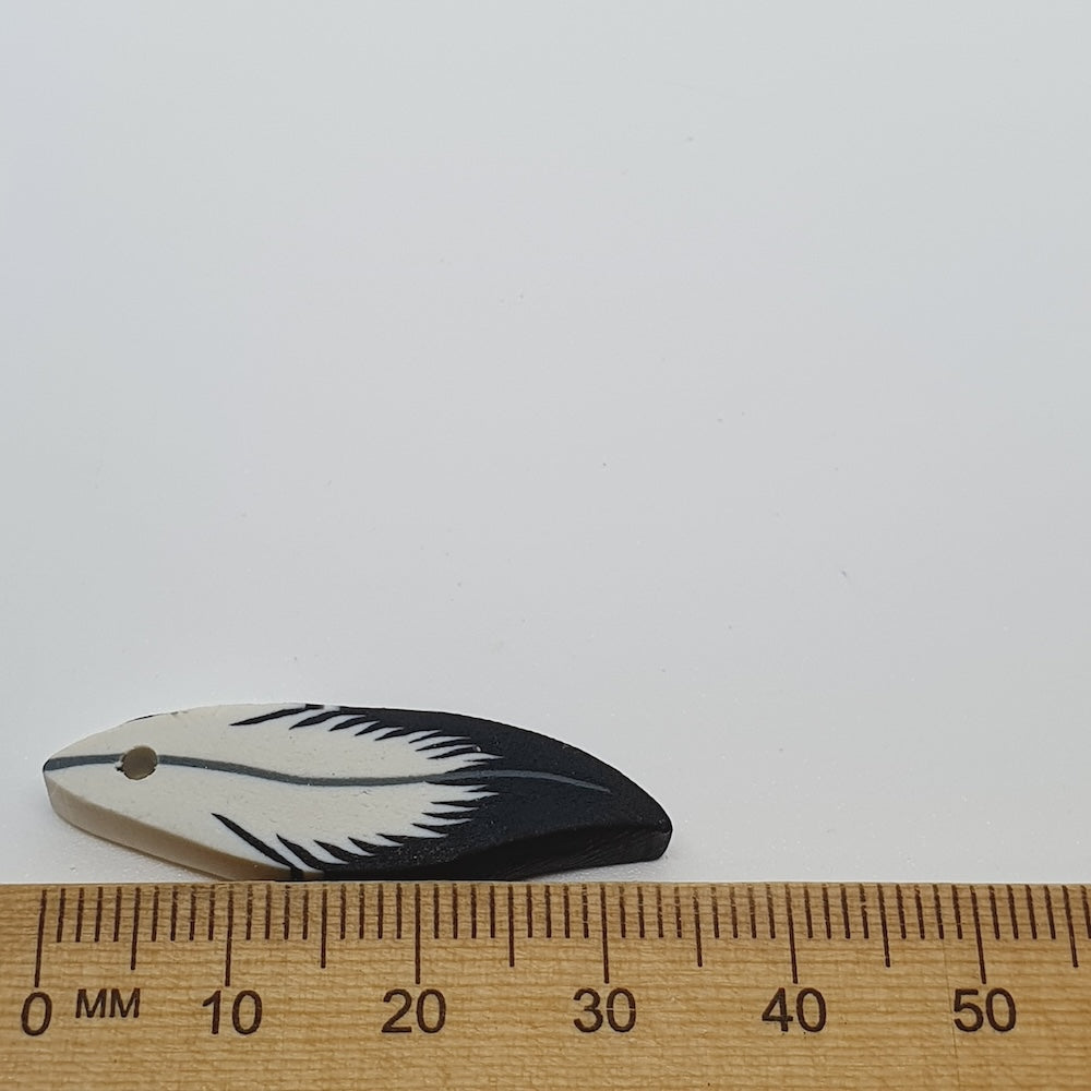 Feather-shaped bead with black and white design on a white background, measured against a ruler.