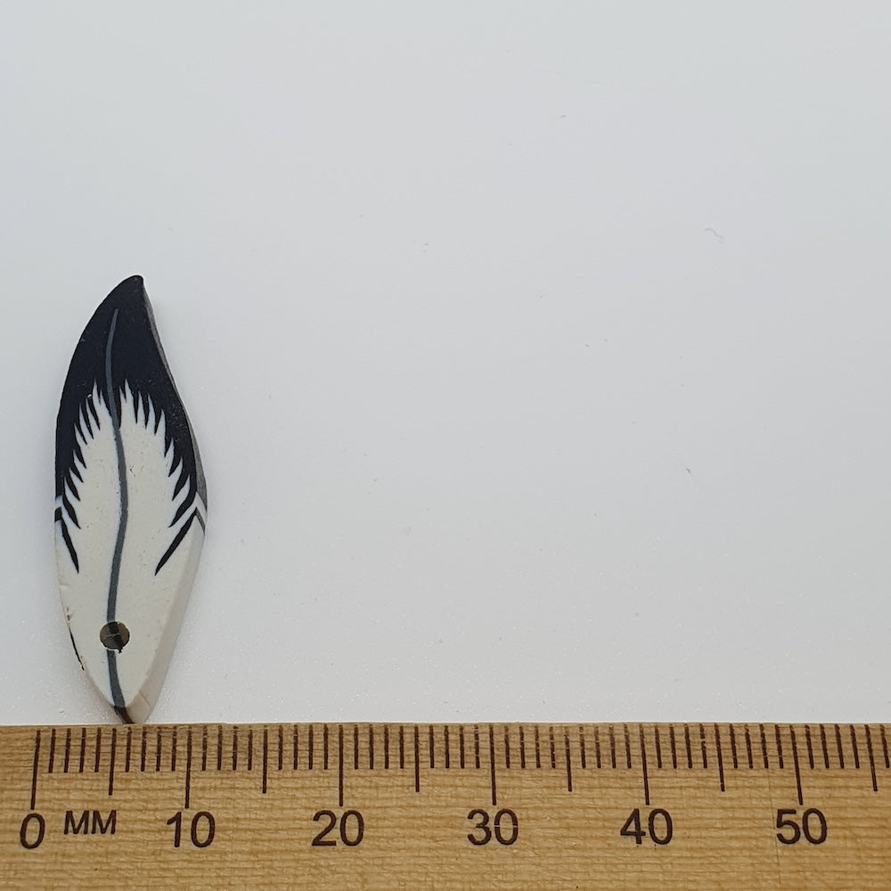 Small feather-shaped bead with black design on a white background, measured against a wooden ruler.