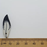 Small feather-shaped bead with black design on a white background, measured against a wooden ruler.