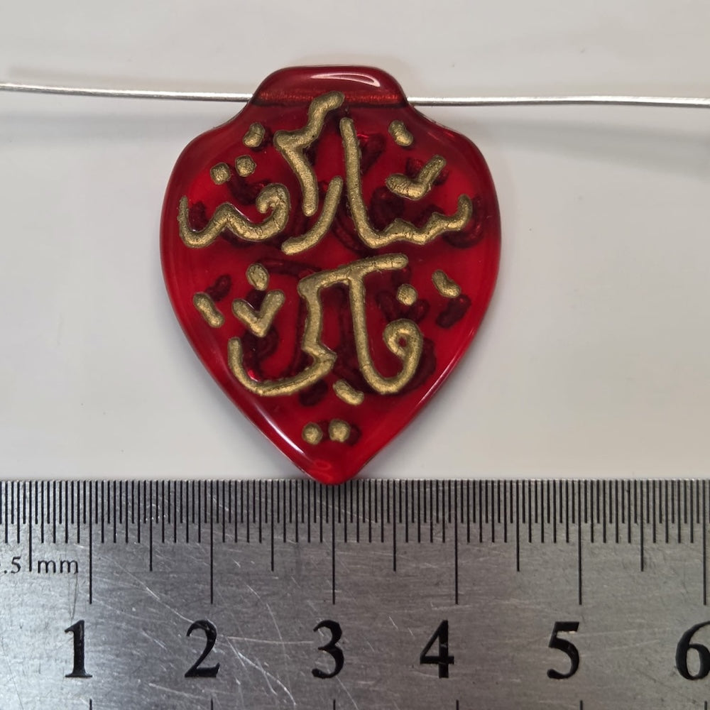 Red pendant with gold Arabic text on a white background with a ruler for scale.