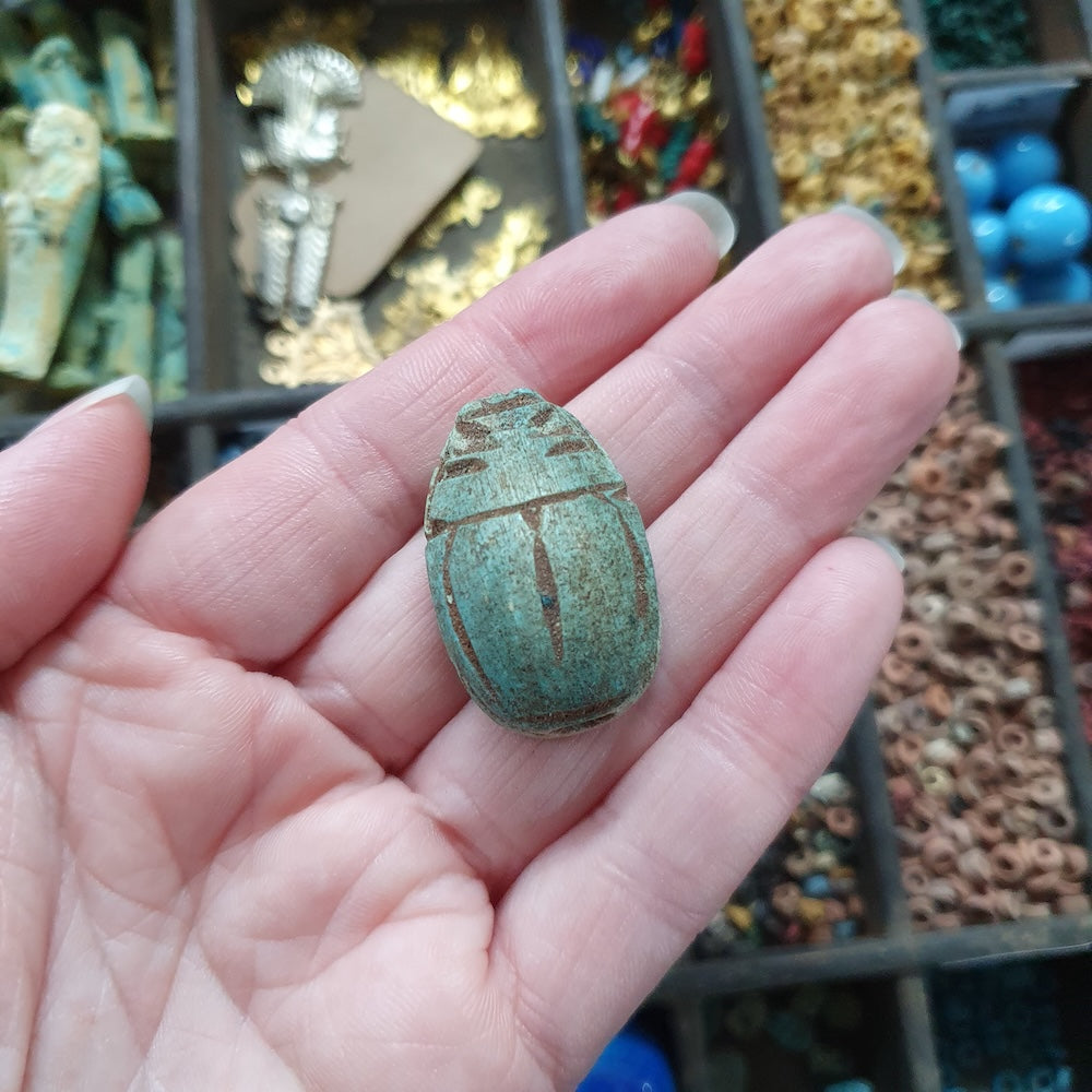 Hand holding a turquoise stone beetle with a blurred background of various stones and items.